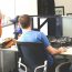 Two people in office, one sitting on chair with two monitors programming