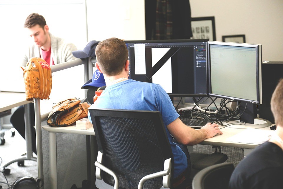 Two people in office, one sitting on chair with two monitors programming